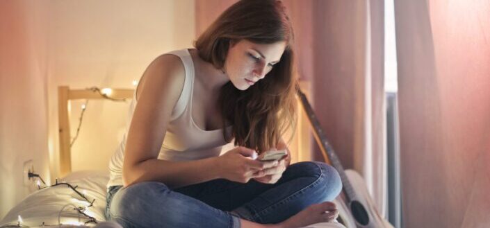 photo-of-woman-in-white-tank-top-and-blue-denim-jeans-sitting-on-bed-while-using-her-phone-